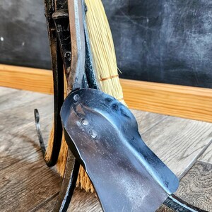 May include: A black metal fireplace tool holder with a broom, shovel, and poker. The tools are hanging on the holder and the broom is resting on the floor.
