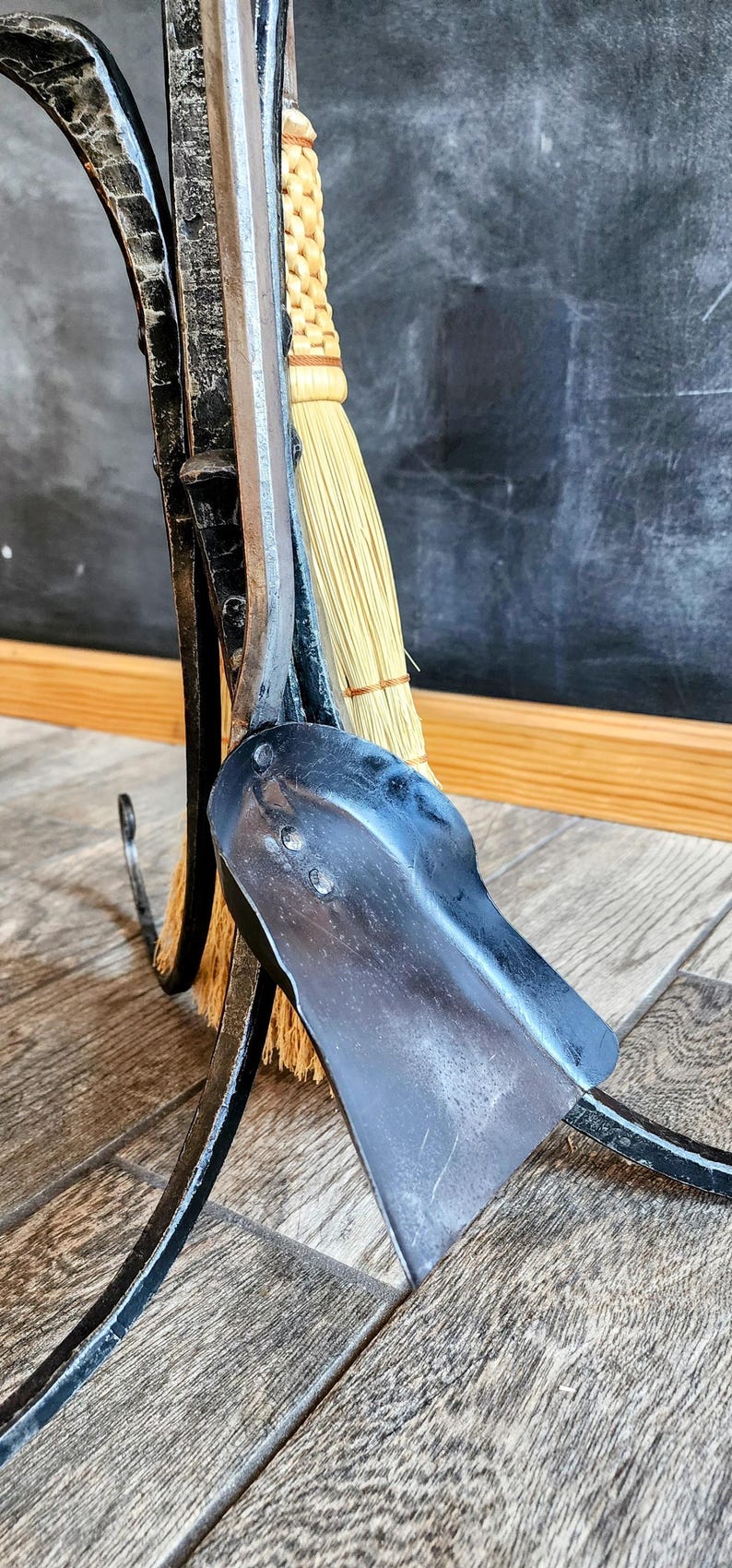 May include: A black metal fireplace tool holder with a broom, shovel, and poker. The tools are hanging on the holder and the broom is resting on the floor.