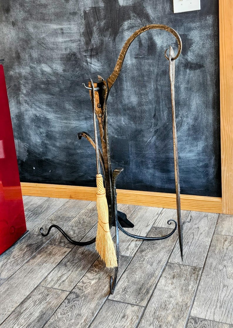 May include: A black metal fireplace tool holder with a curved top and three legs. A long-handled brush and two metal tools are hanging from the holder.