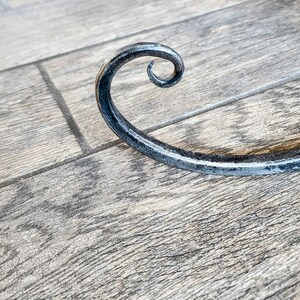 May include: A black metal hook with a decorative curl at the end. The hook is lying on a wooden surface.