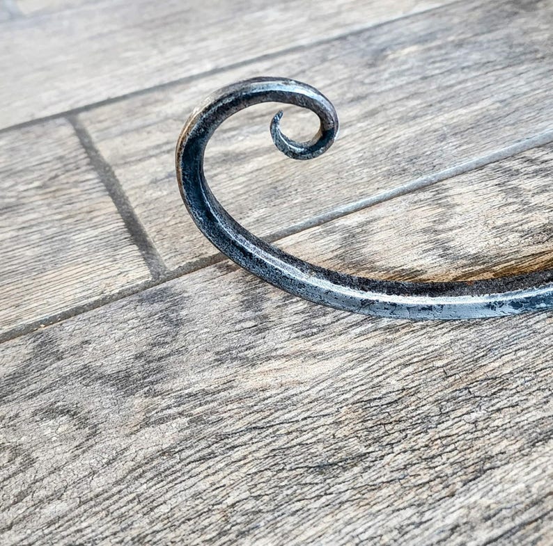 May include: A black metal hook with a decorative curl at the end. The hook is lying on a wooden surface.
