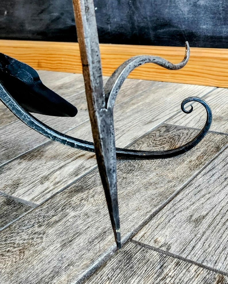 May include: A black metal stand with a decorative scroll design. The stand has a pointed base and a curved top.