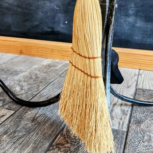 May include: A natural fiber broom with a woven handle hangs on a black metal hook. The broom is made of straw and has a brown twine binding.