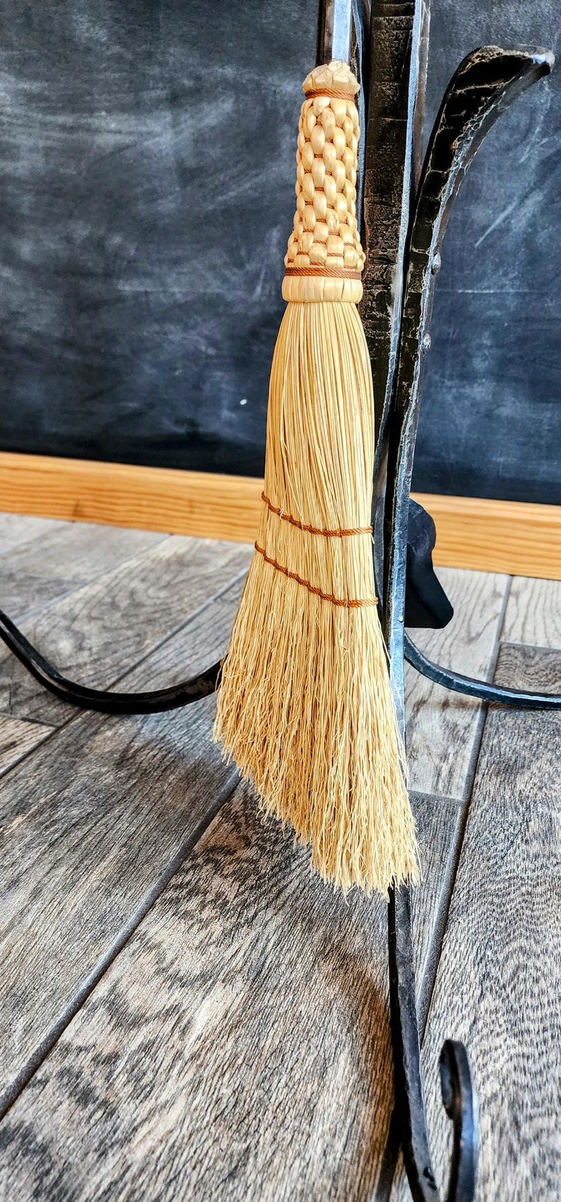 May include: A natural fiber broom with a woven handle hangs on a black metal hook. The broom is made of straw and has a brown twine binding.