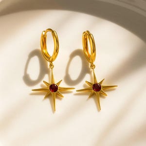 May include: A pair of gold-colored hoop earrings. Each earring features a starburst pendant with a central red gemstone. The earrings are displayed on a white surface, with shadows adding depth.