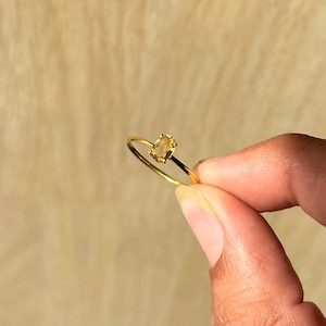 Peut inclure: Une bague en or ornée d'une petite pierre précieuse ovale jaune clair. La monture de la bague est fine et délicate. La bague est tenue par les doigts d'une personne sur un fond flou brun clair.