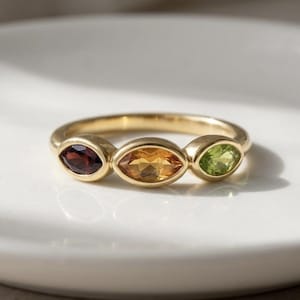 Op de afbeelding: Een gouden ring met drie ovale edelstenen: een dieprode, een goudoranje en een lichtgroene steen. De ring wordt getoond op een wit oppervlak. De edelstenen zijn in een rij geplaatst.
