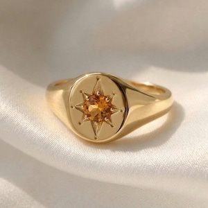 Può includere: Un anello con sigillo dorato con un design a stella e una gemma arancione rotonda centrale. L'anello ha una finitura lucida ed è posto su uno sfondo bianco strutturato. La fascia dell'anello è liscia e arrotondata.