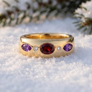 May include: A gold ring with a wide band, featuring an oval garnet gemstone in the centre, flanked by two marquise-cut amethyst gemstones and two small diamonds. The ring is set against a snowy background.