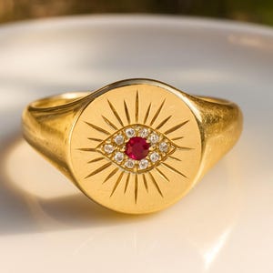 May include: A gold signet ring with an engraved eye design, a central red gemstone, and surrounding white gemstones. The ring has a polished finish and a classic, rounded shape.