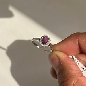 May include: A silver ring featuring a large oval, purplish-red gemstone, surrounded by a halo of smaller, clear stones. The ring is held up against a white background, showcasing its elegant design and sparkle.