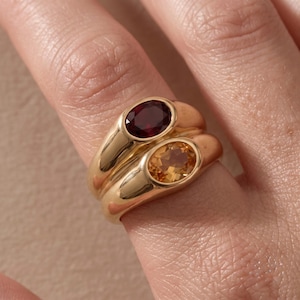 Puede incluir: Dos anillos de oro apilados, cada uno con una banda lisa y pulida. El anillo superior está engastado con una piedra preciosa de corte ovalado de color rojo intenso, mientras que el inferior contiene una piedra ovalada de color amarillo brillante y translúcido.