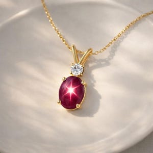 Könnte beinhalten: Eine goldene Halskette mit einem Anhänger, der einen großen, ovalen, tiefroten Edelstein mit Sterneneffekt zeigt. Über dem roten Stein befindet sich ein kleinerer, runder, klarer Edelstein. Die Halskette liegt auf einer weißen Oberfläche.