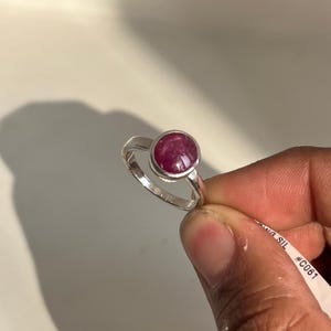 Könnte beinhalten: Ein Silberring mit einem großen, runden, polierten Rubin-Edelstein. Der Rubin hat eine tiefe, satte rote Farbe und ist in einem schlichten, eleganten Silberband gefasst. Der Ring wird von einer Person gehalten.
