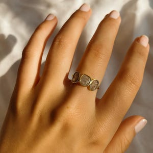 May include: A gold ring with three irregularly shaped, light-coloured stones. The ring is set on a finger, highlighting the design and the warm tones of the metal and stones. The stones have a slightly translucent appearance.