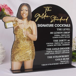 May include: A black acrylic sign with gold lettering displaying "The Golden Standard Signature Cocktails." The sign features a cutout of a woman in a gold dress pouring a drink. A martini glass and floral arrangement are also present.