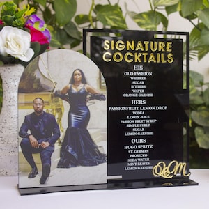 Puede incluir: Un letrero acrílico negro con letras doradas muestra recetas de cócteles. A la izquierda hay una foto de una pareja. A la derecha, una copa de Martini con un cóctel. Arreglos florales rodean el letrero. El letrero dice "SIGNATURE COCKTAILS".