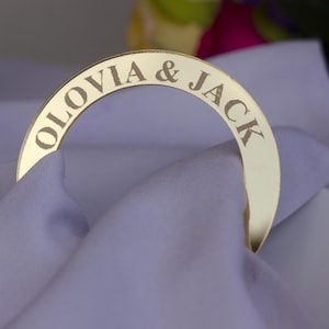 Op de afbeelding: Een goudkleurige servetring met de namen "OLOVIA & JACK" gegraveerd. De ring is gebogen en rust op een gevouwen witte servet. De achtergrond toont een vage bloemstuk.