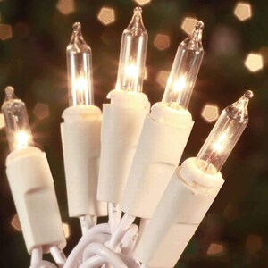 May include: Close-up of a string of clear mini Christmas lights with white bases and wiring. The lights are illuminated, emitting a warm, golden glow. The background is blurred, with hints of bokeh.