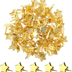 Puede incluir: Un conjunto de chinchetas doradas en forma de estrella. Las chinchetas tienen un acabado metálico brillante y están dispersas por toda la imagen. También se ven varias chinchetas individuales en la parte inferior, mostrando su diseño.