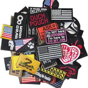 Op de afbeelding: Een verzameling geborduurde patches in verschillende vormen en maten. Veel patches tonen de Amerikaanse vlag, samen met tekst als "OUCH POUCH", "ASK" en "I'm NOT here for your entertainment". Andere patches bevatten een schedel, pootafdrukken en een slang.