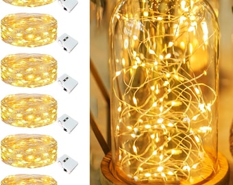 May include: A display of warm white LED string lights, coiled and ready for use, alongside a glass cloche filled with similar lights. The cloche sits on a wooden base, creating a cozy, ambient glow. The lights are ideal for home decor.