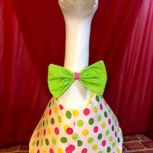 May include: A white goose with a yellow beak and a green bow tie with a pink center. The goose is wearing a white vest with colorful polka dots in yellow, green, and pink. The vest has pink buttons.