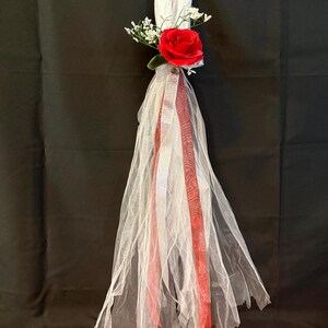 May include: A decorative hanging ornament featuring a cluster of white tulle fabric gathered at the top. A single vibrant red rose is centered with small white baby's breath flowers. Two long ribbons, one silver and one red, hang down the center.