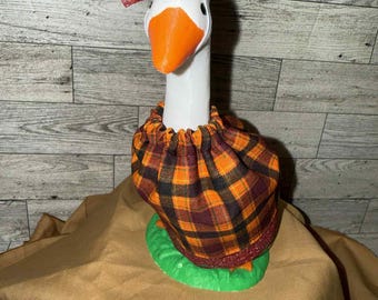 May include: A white goose figurine with an orange beak and a decorative bow. The goose wears a plaid fabric collar and sits on a green base. The bow and collar feature orange, brown, and red hues.