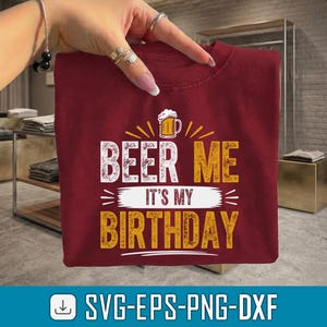 May include: A maroon t-shirt with the text "BEER ME IT'S MY BIRTHDAY" in white and gold. The design includes a beer mug graphic. The shirt is folded and held up by a hand.