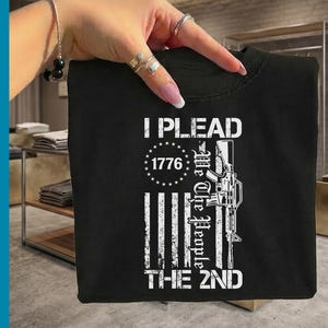 Op de afbeelding: Zwart T-shirt met witte tekst en afbeeldingen. Het ontwerp bevat de woorden "I PLEAD" boven "1776" in een cirkel van sterren. Daaronder staat "We The People THE 2ND" met een geweer afbeelding.