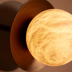 以下が含まれることがあります： モダンなウォールスコンス。 大きな質感のある球形の照明器具が特徴です。 光は暖かく輝く黄色で、ブロンズ色の円形バックプレートにセットされています。 この器具は、暗く細いアームに取り付けられており、印象的なコントラストを生み出しています。