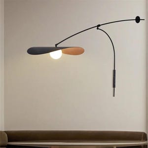 Può includere: Una moderna lampada da parete con un braccio nero curvo e un paralume grigio scuro e beige. La lampada ha una lampadina sferica ed è progettata per l'illuminazione interna. Il braccio si estende da un supporto a parete.