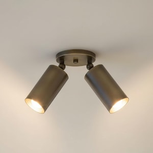 Könnte beinhalten: Ein bronzefarbener Deckenstrahler mit zwei verstellbaren zylindrischen Lichtern. Die Lichter sind nach unten gerichtet und strahlen ein warmes Licht aus. Die Leuchte ist auf einer kreisförmigen Basis montiert.