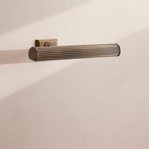 May include: A bronze-colored, cylindrical wall-mounted light fixture. The fixture has a rectangular base and a ribbed, horizontal light bar. The light is mounted on a light-colored wall.