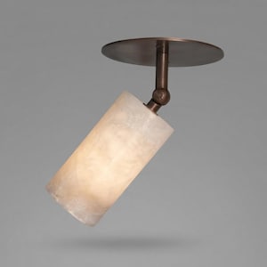 May include: A ceiling-mounted spotlight featuring a circular dark bronze base and a cylindrical, translucent, off-white stone shade. The fixture includes an adjustable swivel joint connecting the base to the light housing, allowing for directional lighting.