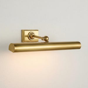 May include: A brass-colored picture light fixture. The fixture has a rectangular base with a curved arm and a cylindrical light bar. The light bar is positioned horizontally, and the fixture is mounted on a white wall.