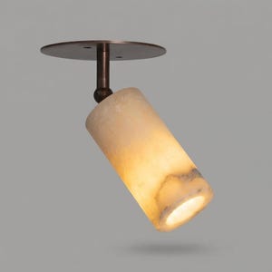 May include: A ceiling-mounted spotlight featuring a cylindrical shade made of translucent, cream-colored stone with natural veining. The fixture includes a dark bronze-toned metal base and an adjustable swivel arm that connects the shade to the mount.