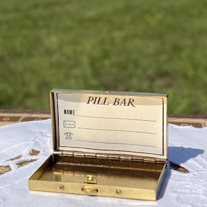 Op de afbeelding: Een goudkleurig metalen pillendoosje met een scharnierend deksel. De binnenkant van het deksel bevat de tekst PILL BAR en een gedrukt formulier met lijnen voor een naam, een envelop-icoon en een telefoon-icoon. Het doosje heeft een eenvoudige sluiting.