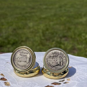 Könnte beinhalten: Zwei runde goldfarbene Metalldosen mit Klappdeckeln. Jeder Deckel hat eine kreisförmige silberfarbene Relief-Einlage, die ein Gebäude darstellt. Die linke Einlage zeigt La Poma und die rechte Einlage zeigt Casa de la Vall Andorra.