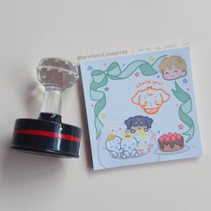 May include: A black self-inking stamp with a clear diamond-shaped handle next to a square memo sheet featuring a light green ribbon border, a cartoon dog, a chocolate cake with strawberries, and the text @harmlesslynapping and thank you.