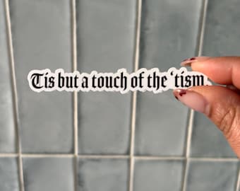May include: A rectangular white sticker with a black border and black text in a gothic font that reads, Tis but a touch of the 'tism. The sticker has a die-cut shape that follows the contour of the lettering, creating a clean and bold visual appearance.