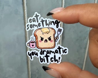 Puede incluir: Una pegatina troquelada con una rebanada de pan tostado de dibujos animados con mantequilla, expresión facial enfadada, sosteniendo una taza rosa y un tenedor. El texto dice eat something you dramatic bitch! en letras negras estilizadas.