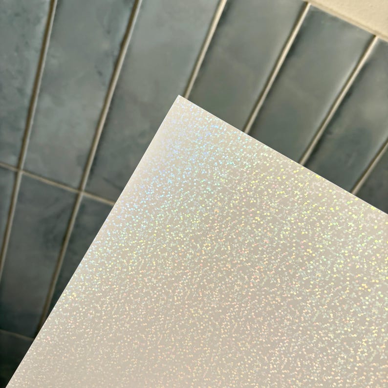 May include: A rectangular sheet of white material featuring a fine, holographic glitter finish that reflects light in a spectrum of colors. The surface has a dense, uniform texture of tiny sparkling particles across the entire visible area.