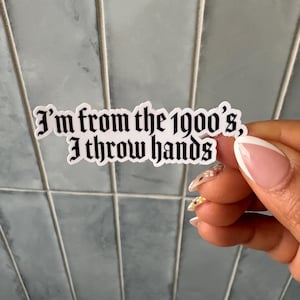 Puede incluir: Una pegatina blanca con el texto "I'm from the 1900's, I throw hands" en una fuente gótica. La pegatina se sostiene contra un fondo de azulejos azules. La pegatina es rectangular y tiene un acabado brillante.