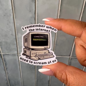 Könnte beinhalten: Ein Aufkleber mit einem Retro-Computer und dem Text "I remember when the internet used to scream at us." Der Computerbildschirm zeigt "CONNECTING..." mit einem Ladebalken. Der Aufkleber wird von einer Person gehalten.