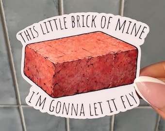 Puede incluir: Una pegatina troquelada con un bloque rectangular texturizado de color rosa rojizo que recuerda a la carne procesada. El texto "THIS LITTLE BRICK OF MINE" está impreso en negro arriba y "I'M GONNA LET IT FLY" en negro abajo.