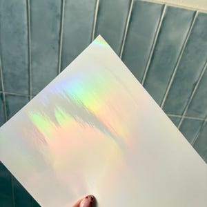 May include: A rectangular sheet of white material featuring a prominent holographic finish that reflects a spectrum of iridescent colors including green, yellow, and pink across its surface, creating a light-catching effect on the smooth texture.