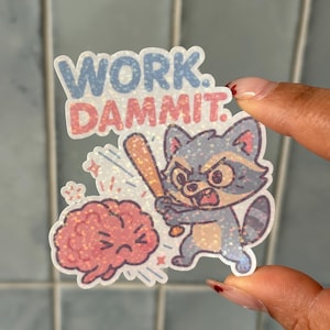 Op de afbeelding: Een sticker met de tekst "WORK. DAMMIT." boven een illustratie van een boze wasbeer met een honkbalknuppel, op het punt om een hersen te slaan. De sticker heeft een glitterafwerking en een cartoonstijl.