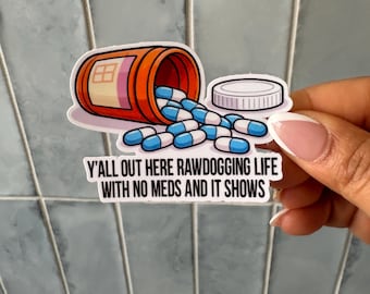 May include: A die-cut sticker featuring an orange pill bottle tipped on its side with blue and white capsules spilling out, accompanied by a white bottle cap. The text reads: Y'ALL OUT HERE RAWDOGGING LIFE WITH NO MEDS AND IT SHOWS.